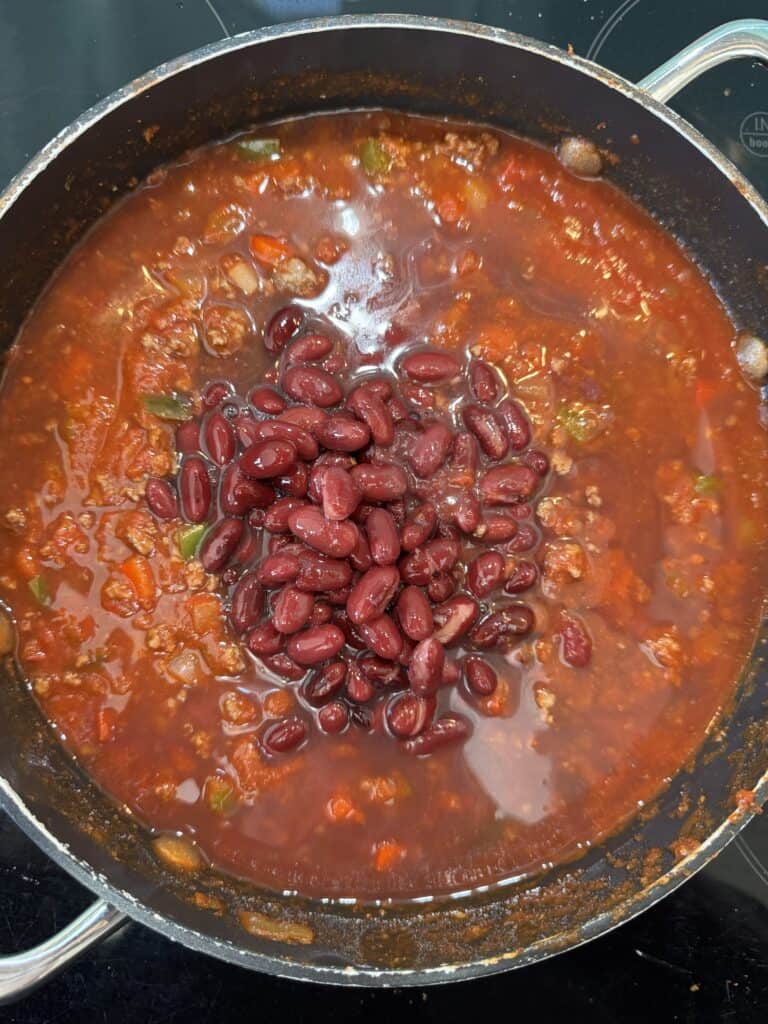 A pot filled with chili, containing tomatoes, ground meat, and a mix of red kidney beans and other vegetables. The chili is simmering, with kidney beans prominently displayed on top.
