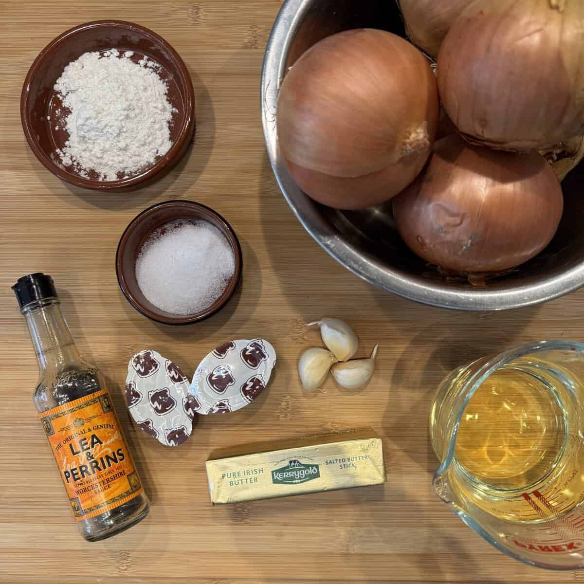 Ingredients on a wooden surface include a bottle of Worcestershire sauce, a stick of Kerrygold butter, peeled garlic cloves, a bowl of onions, a bowl of flour, a bowl of salt, three beef stock cubes, and a measuring cup of broth.