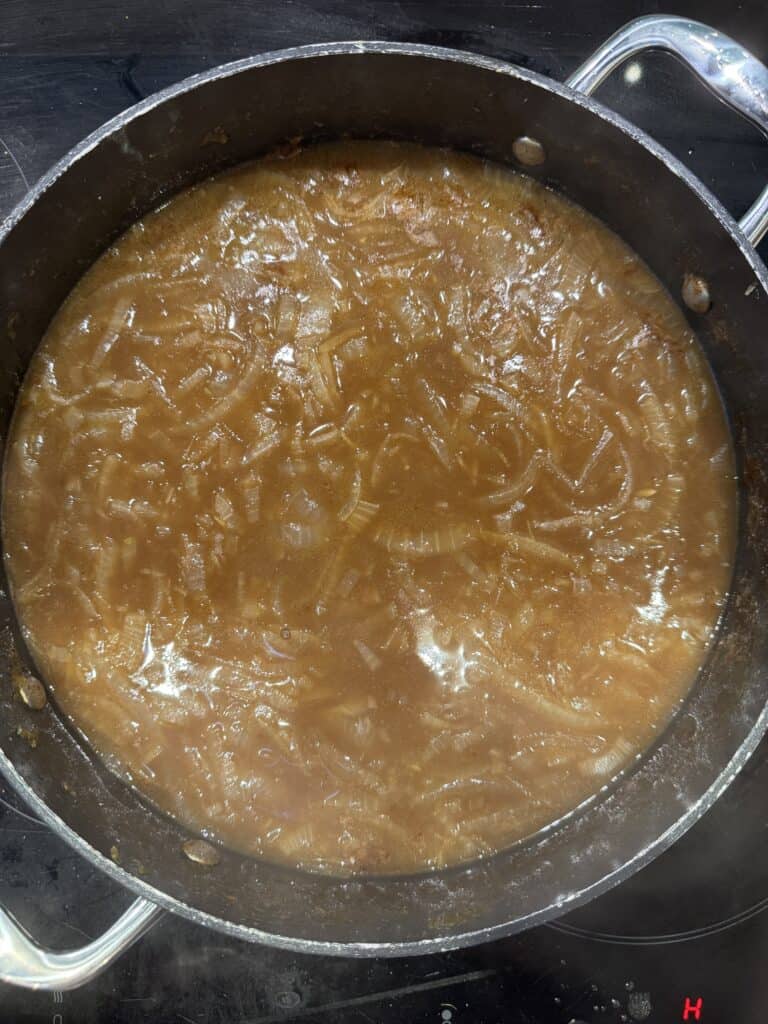 A pot filled with steaming French onion soup, featuring translucent sliced onions in a rich, brown broth. The pot sits on a black stovetop with two handles visible on either side.