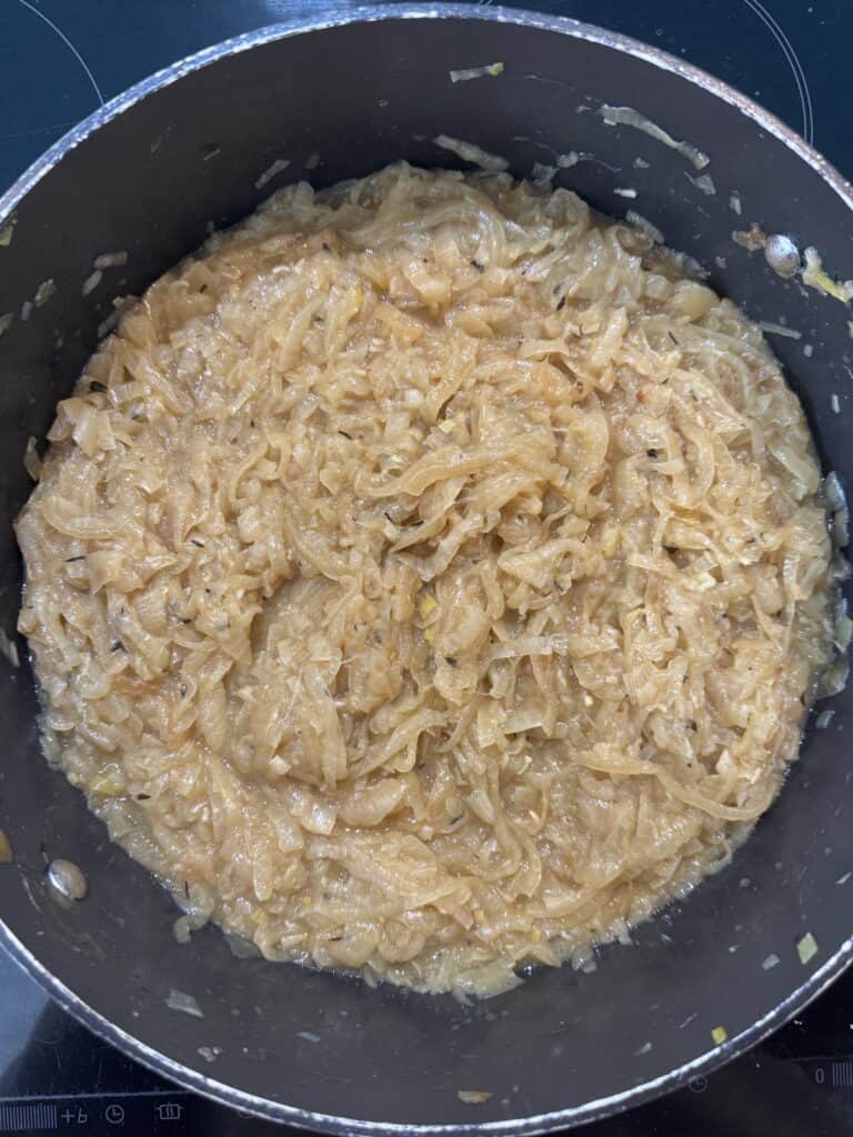 A pot of caramelized onions, cooked to a soft, golden-brown color, filling the bottom of a black saucepan on a stovetop. The onions appear tender and evenly browned.