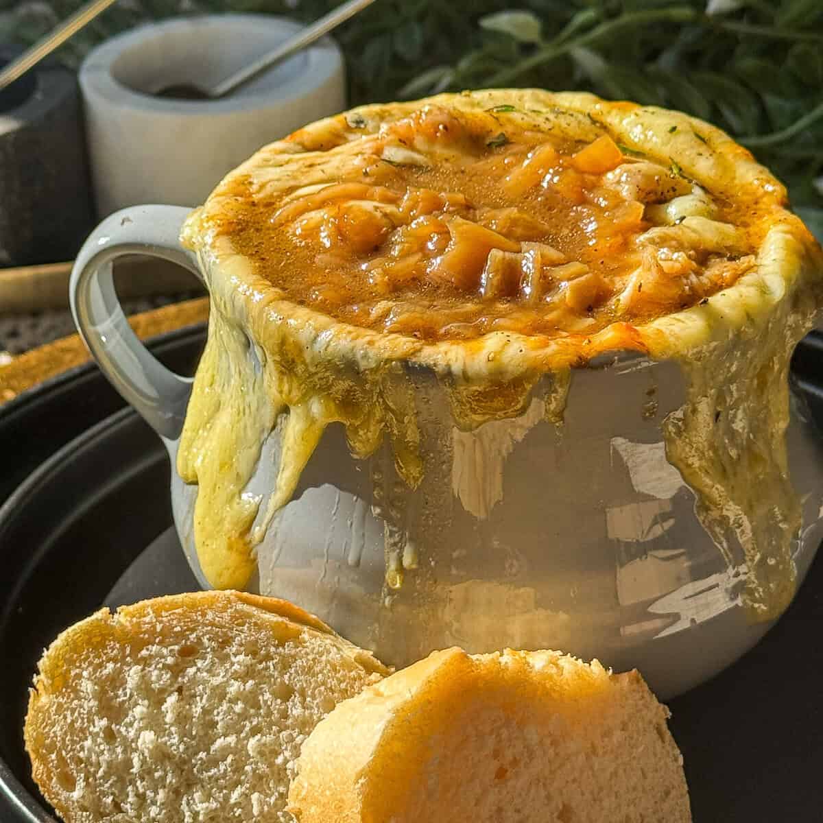 A mug of French onion soup is topped with melted cheese, overflowing down the sides. Two slices of bread are beside the mug on a dark plate. The background includes a blurred plant and kitchen items.