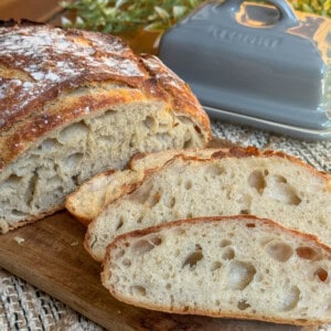 Close-up of freshly baked, sliced rustic bread on a wooden board. The bread has a golden, crusty exterior and a soft, airy interior. A gray butter dish with a lid is in the background, with some greenery nearby.