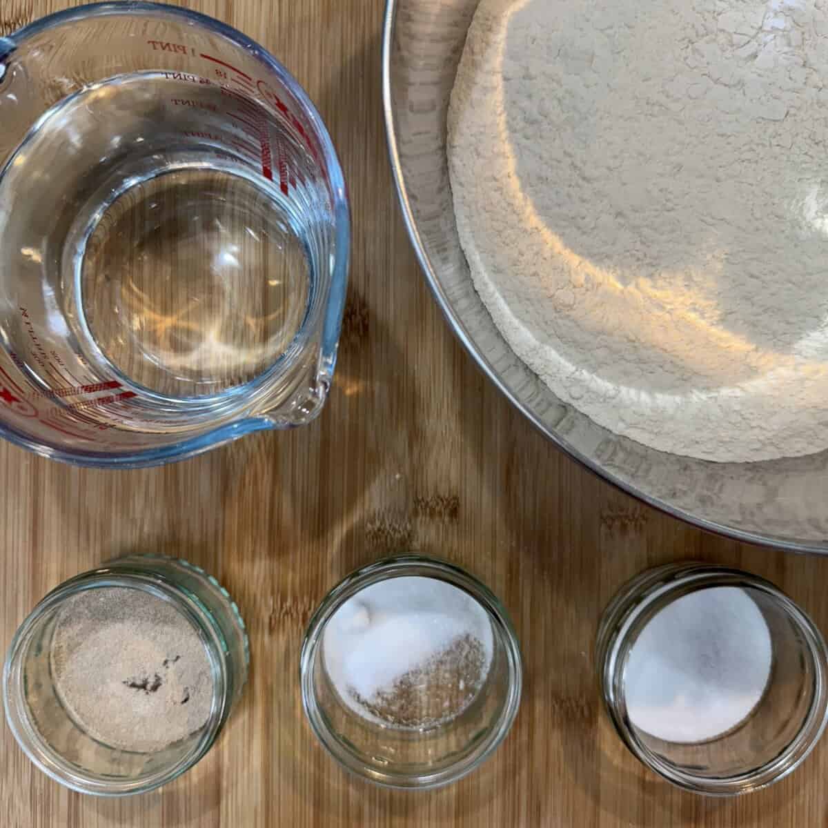 A top-down view of a clear measuring cup with water, a metal bowl with flour, and three small glass jars containing yeast, salt, and sugar on a wooden surface.