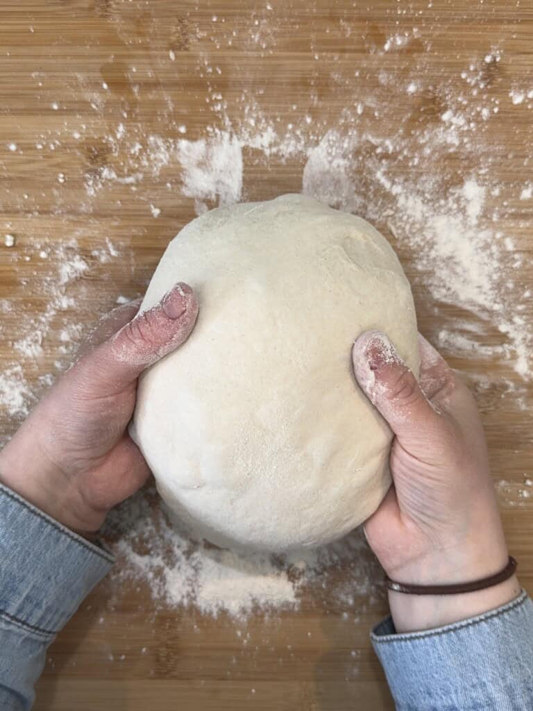Hands holding a round, smooth dough ball on a floured wooden surface. The person is wearing a light-colored, long-sleeved shirt. Flour is scattered around.