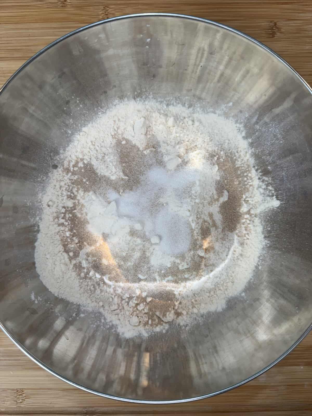 A stainless steel bowl containing flour, sugar, and yeast seen from above on a wooden surface. The ingredients are dry and not yet mixed, forming distinct layers within the bowl.