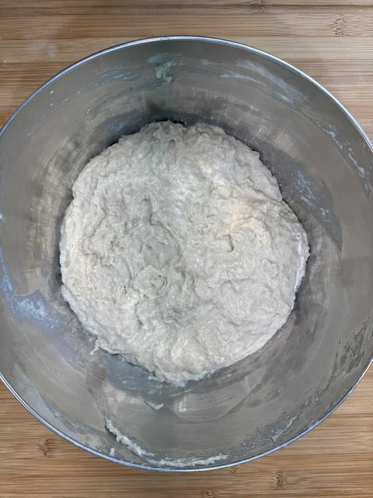 A metal mixing bowl filled with a lumpy, wet dough sits on a wooden surface. The dough appears to be in the early stages of mixing, with a rough texture and uneven consistency.