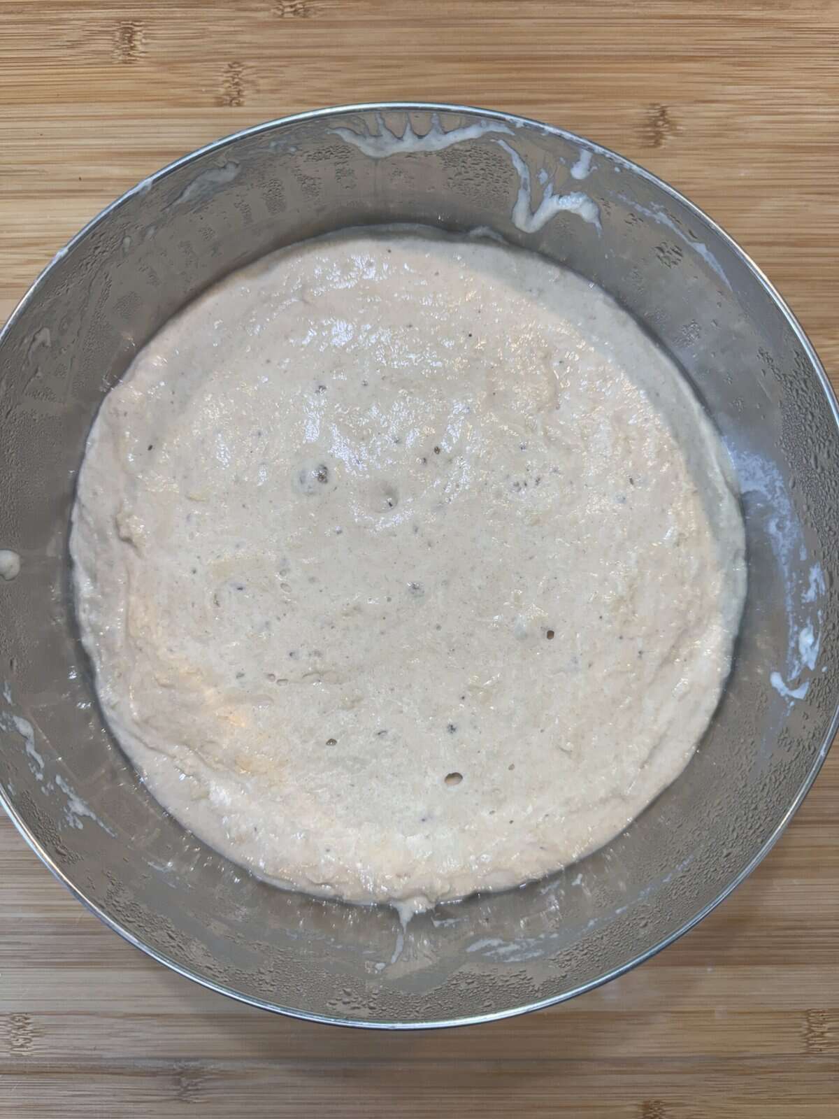 A metal mixing bowl filled with a creamy, light tan batter is placed on a wooden surface. The batter appears smooth with small air bubbles on its surface, suggesting its ready to be cooked or baked.