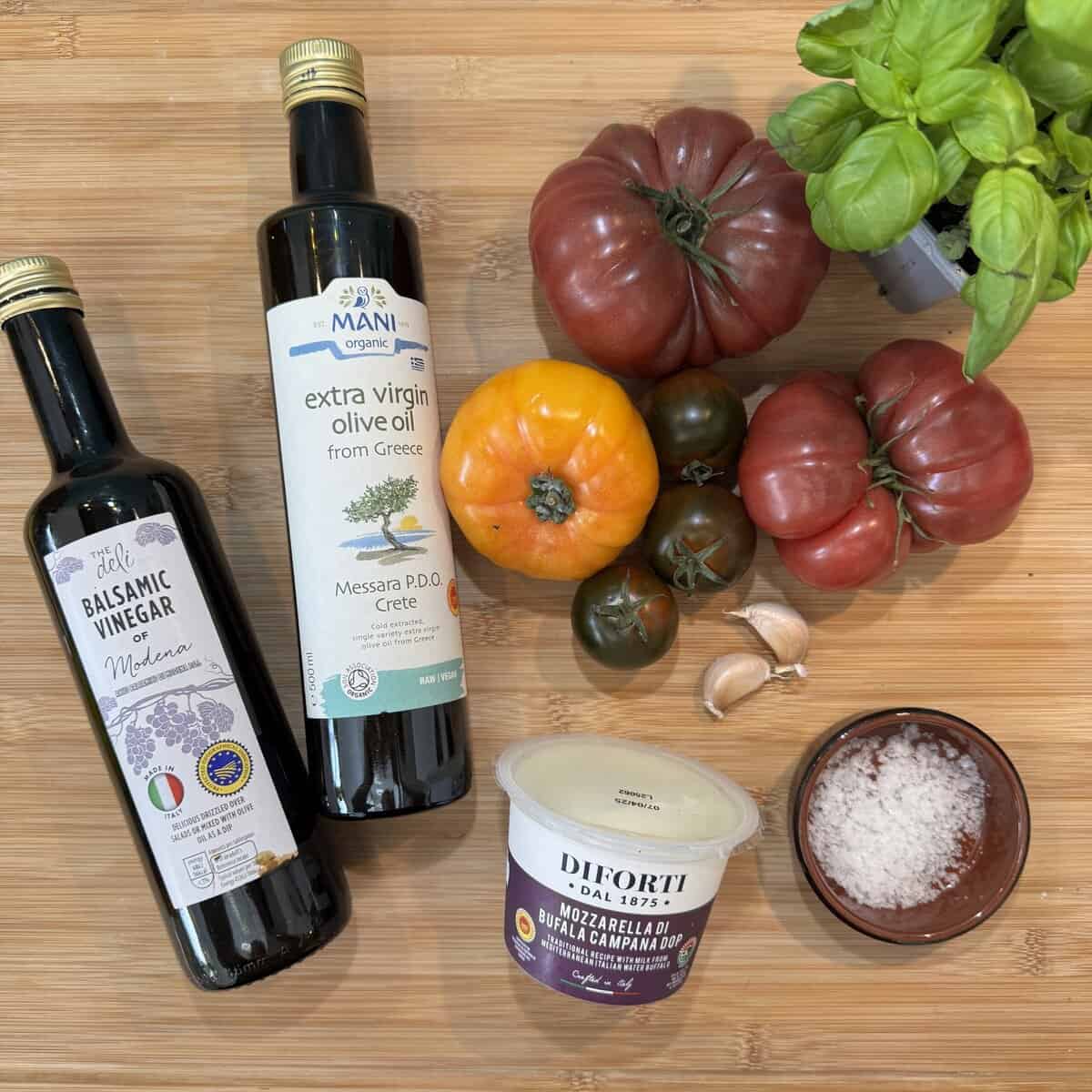 Bottles of balsamic vinegar and extra virgin olive oil are on a wooden surface with heirloom tomatoes, fresh basil, garlic cloves, a bowl of coarse salt, and a container of buffalo mozzarella cheese.
