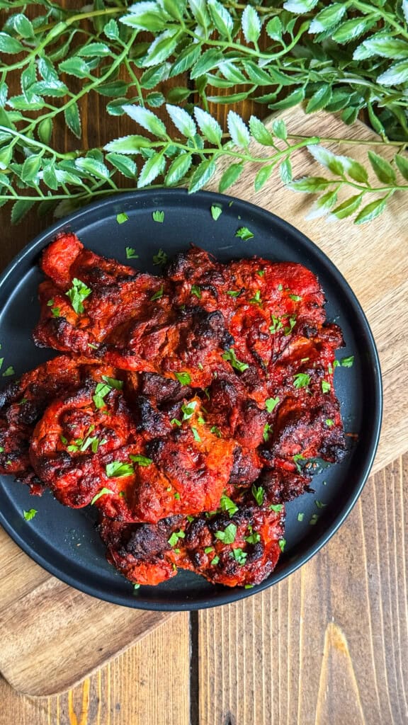 A black plate with pieces of grilled, red-marinated chicken garnished with chopped herbs, placed on a wooden table beside leafy green plants.