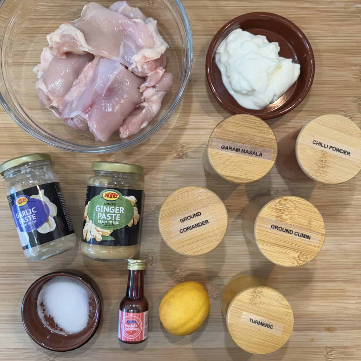 Top-down view of ingredients for chicken marinade: raw chicken, yogurt, jars of garlic and ginger paste, lemon, salt, red food coloring, and spice jars labeled garam masala, chili powder, coriander, cumin, and turmeric.