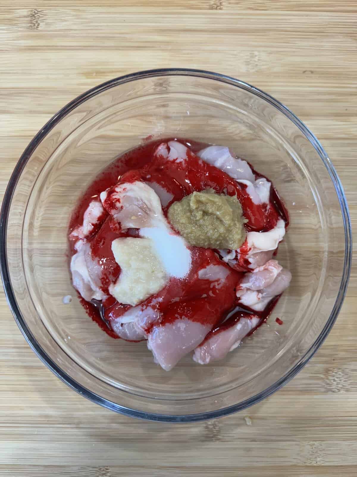 A glass bowl containing raw chicken pieces, red marinade, a dollop of ginger-garlic paste, and a sprinkle of white salt, placed on a wooden surface.