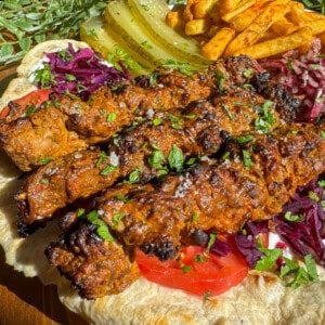 Grilled kebab skewers served on flatbread with sliced tomatoes, purple cabbage, pickles, seasoned fries, and fresh herbs, all garnished with chopped parsley.