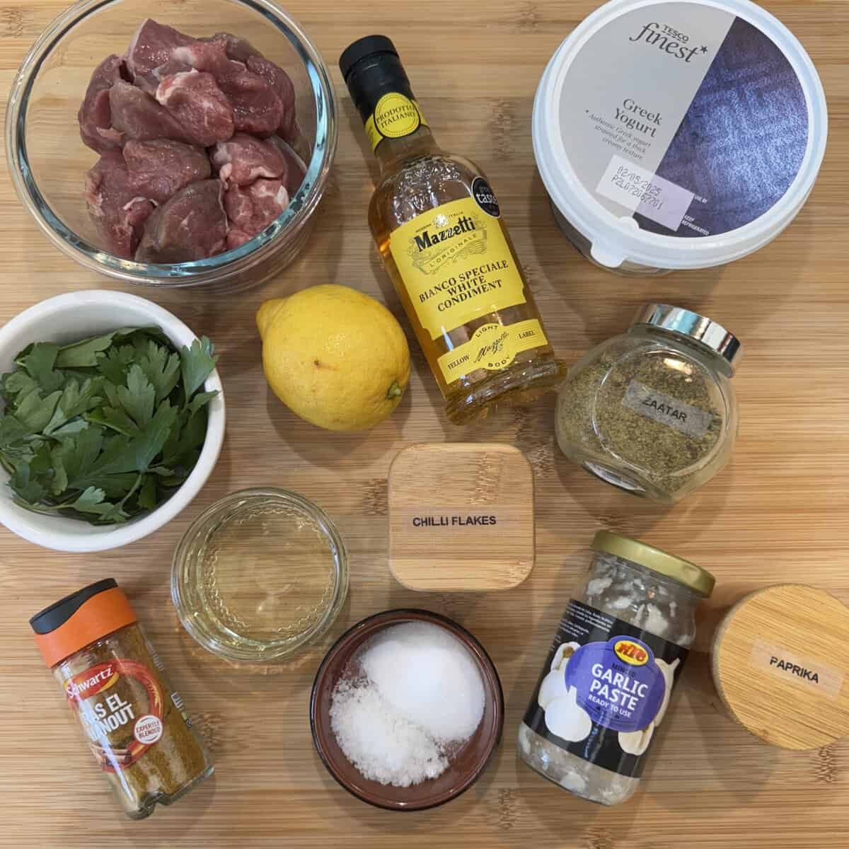 Top-down view of ingredients for  lamb shish kebabs on a wooden surface: raw lamb chunks, Greek yogurt, a bottle of condiment, a lemon, parsley, za’atar, chili flakes, salt, garlic paste, oil, paprika, and salt & pepper seasoning.