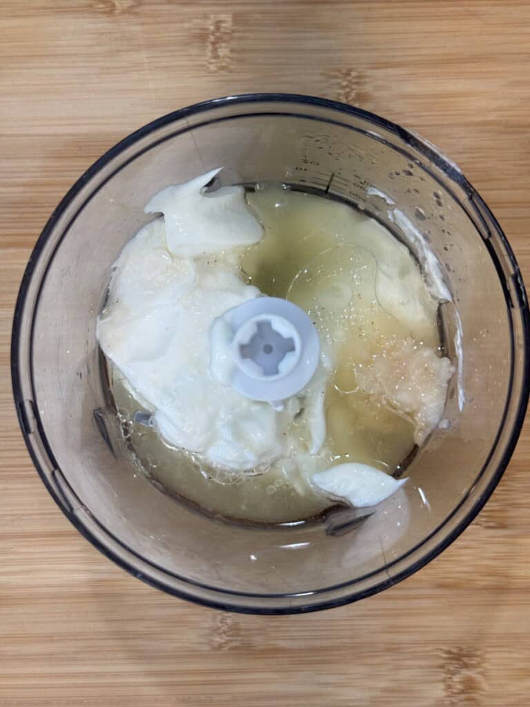 A food processor bowl contains egg whites and a clear liquid, possibly lemon juice or water, sitting on a light wooden surface.