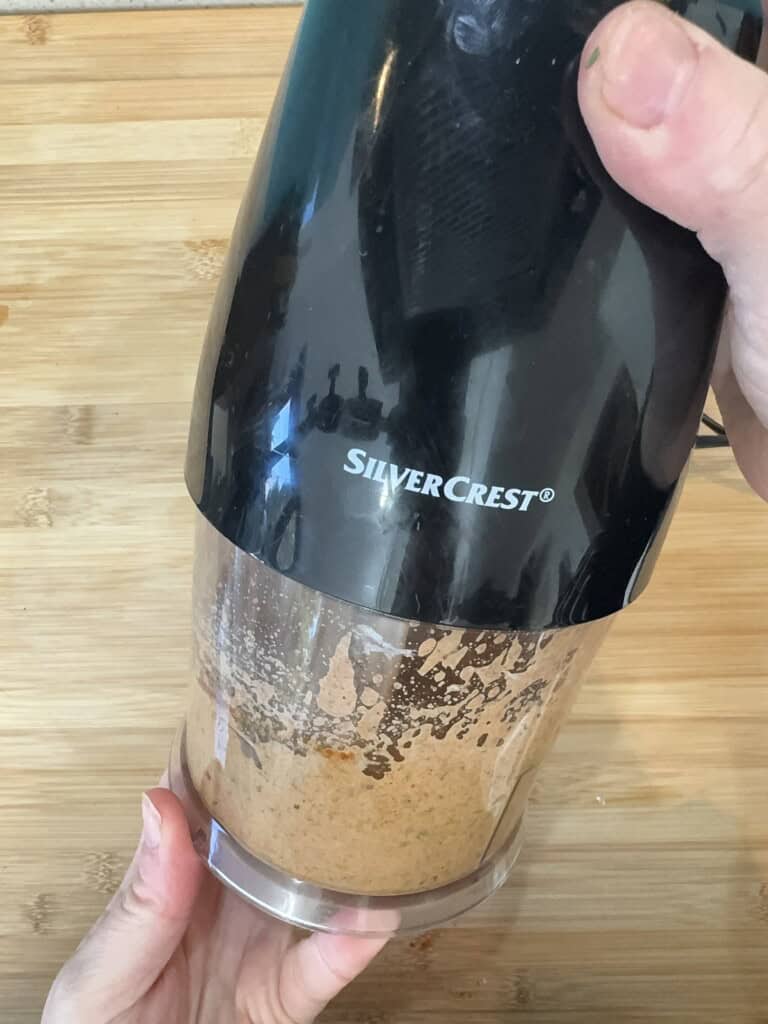 A person holding a SilverCrest electric grinder blending a brown mixture on a wooden surface.