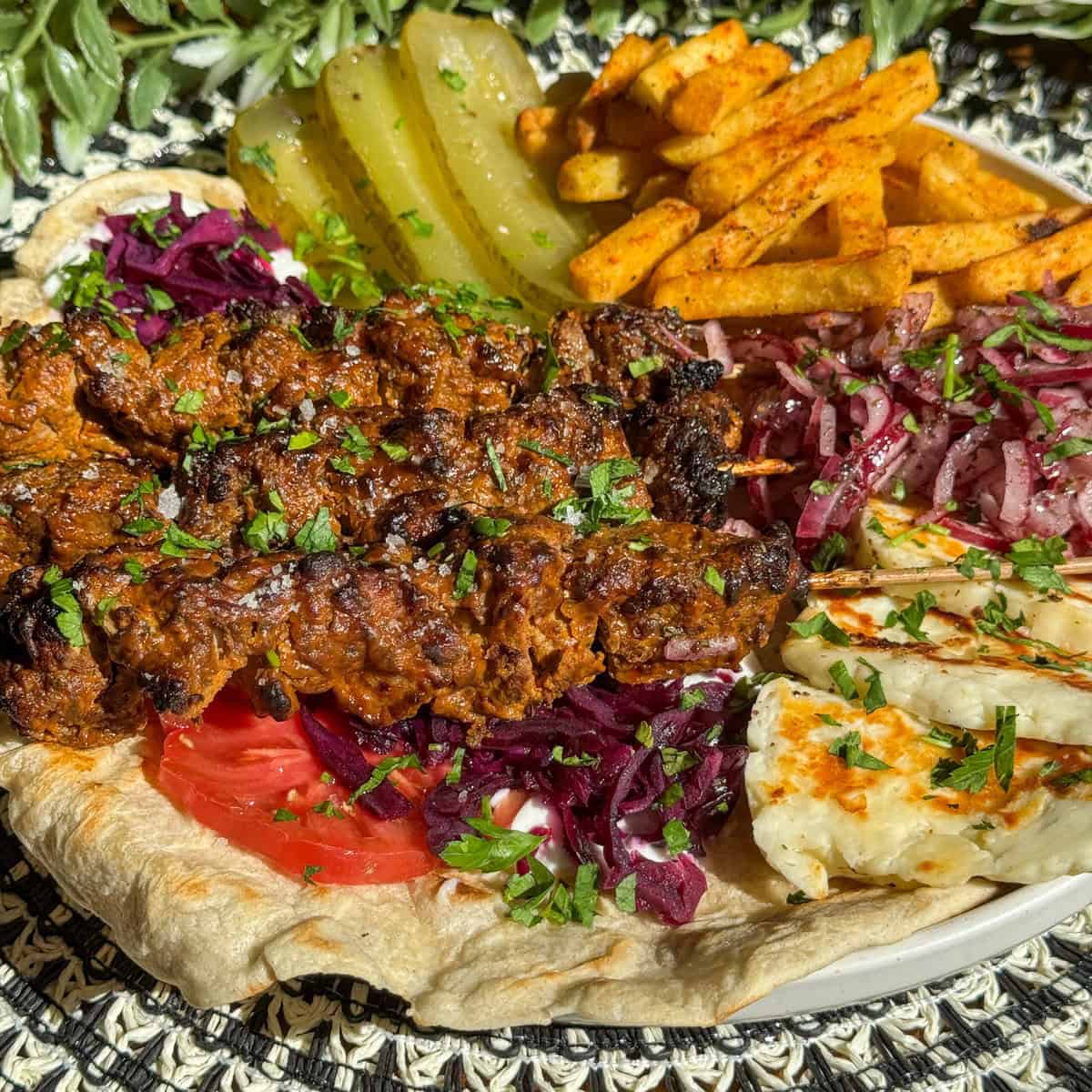 A plate with grilled lamb shish kebab meat skewers on flatbread, sliced pickles, spiced fries, grilled cheese, shredded red cabbage, sliced tomatoes, and red onion, all garnished with chopped herbs.
