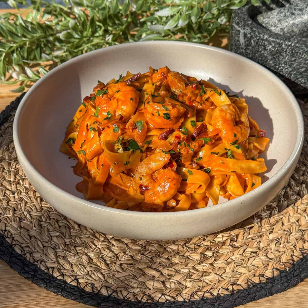 A bowl of tagliatelle pasta with prawns and chorizo in a rich tomato sauce, garnished with herbs, sits on a woven placemat with green foliage in the background.
