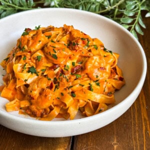 A bowl of creamy tagliatelle pasta with prawns and chorizo, garnished with chopped parsley, sits on a wooden table with green leafy decorations in the background.