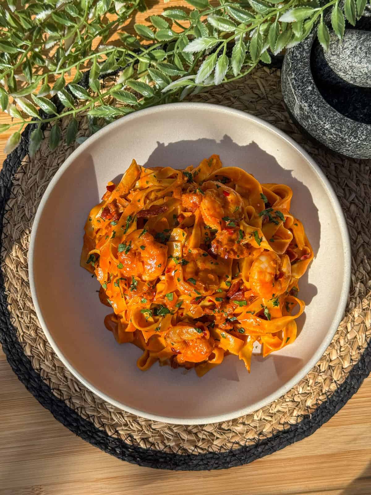 A bowl of tagliatelle pasta topped with prawns and chorizo in a rich tomato sauce, garnished with herbs, is placed on a woven mat. Sunlight highlights the dish, and green leaves are partly visible above.