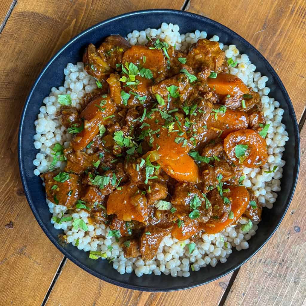 A black bowl filled with couscous topped with a rich tagine of beef, carrots, and herbs, garnished with chopped parsley, sits on a wooden table.