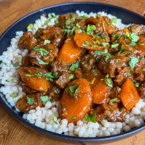 A black bowl filled with pearl couscous topped with a hearty beef and carrot stew, garnished with fresh chopped parsley. The dish is set on a wooden surface.