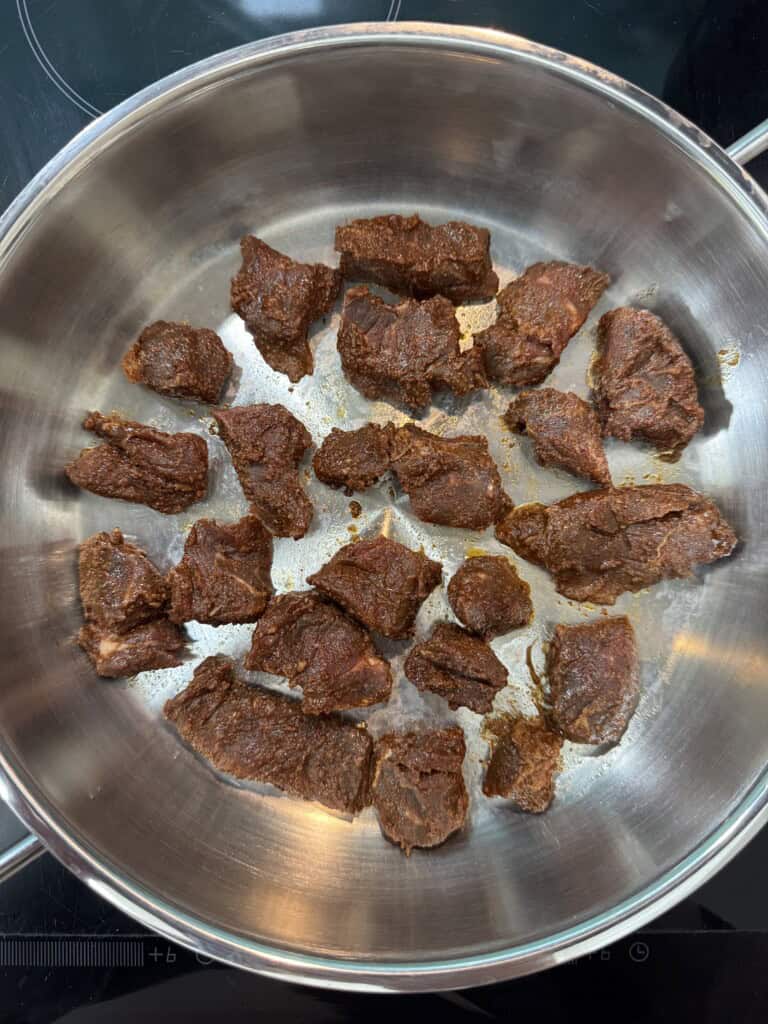 Chunks of seasoned beef are browning in a large stainless steel pan on a stovetop, with some pieces showing a slightly seared exterior.