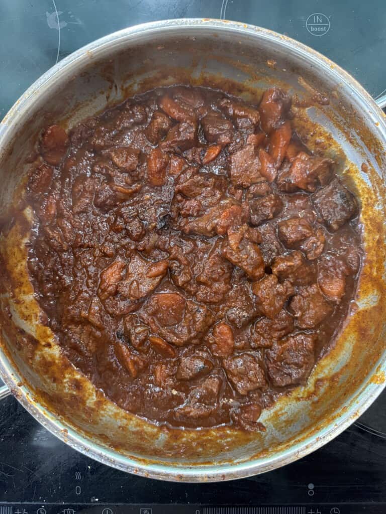 A pot of beef stew simmers on a stovetop, with chunks of beef and carrots in a thick, rich brown sauce.