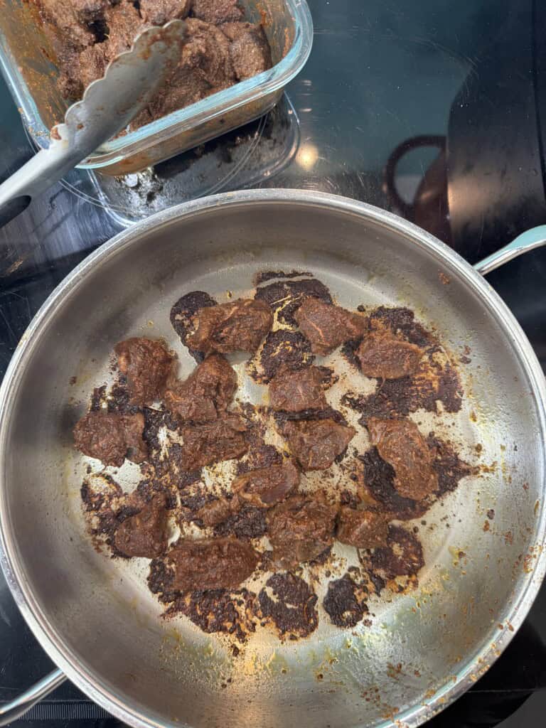 A stainless steel pan on a stovetop with browned and slightly burnt chunks of marinated meat. A pair of tongs rests in a glass container with more marinated meat in the background.