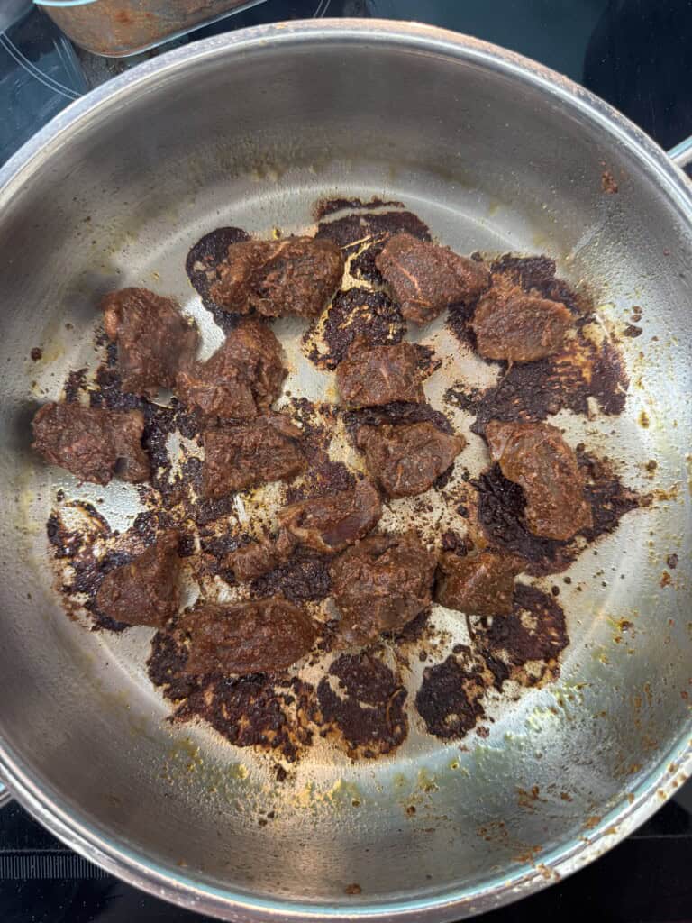 Slices of food with a dark brown, almost burnt exterior are scattered in a stainless steel pan on a stovetop, showing signs of being cooked or seared.