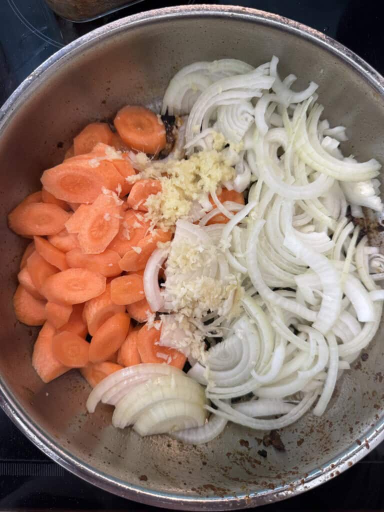 A pot filled with sliced carrots, sliced onions, and minced garlic, ready to be cooked. The vegetables are uncooked, and the pot shows some browning at the bottom.