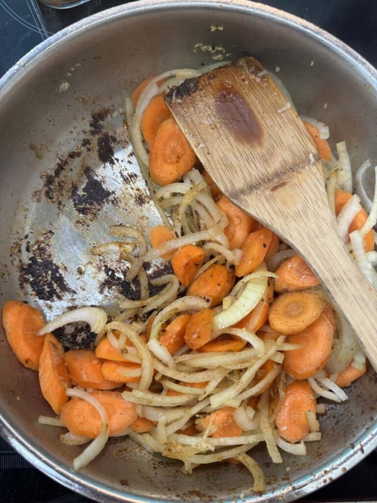 Sliced carrots and onions are being sautéed in a pan with some browning on the bottom. A wooden spatula rests on top of the vegetables.