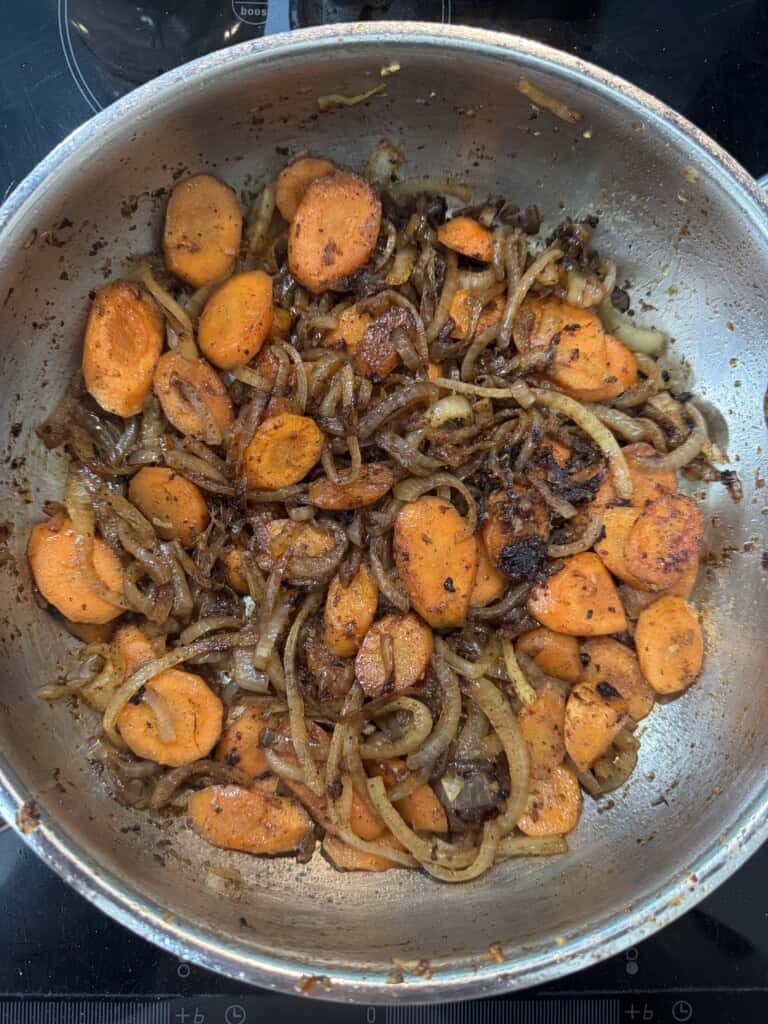 Sliced carrots and onions are sautéing in a pan, showing a mix of caramelized and slightly browned pieces on a stovetop.