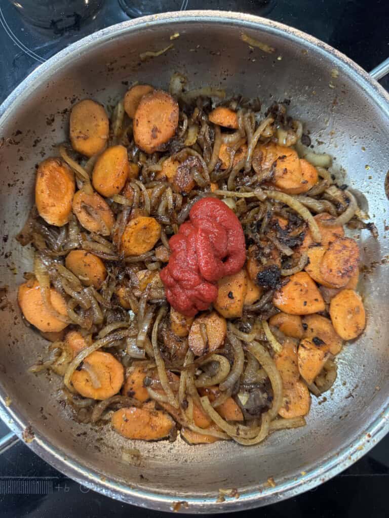 Sliced carrots, onions, and browned bits are sautéing in a pan with a dollop of tomato paste in the center, ready to be stirred in.