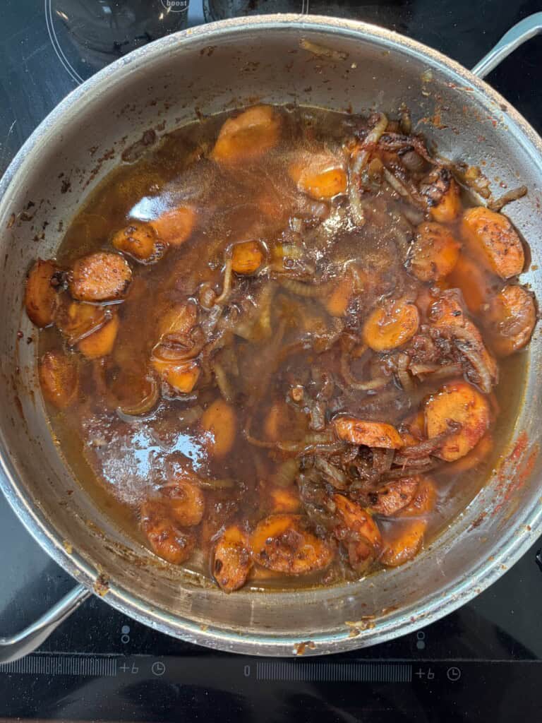 A metal pan on a stovetop filled with a stew of sliced carrots, shredded meat, and onions simmering in a rich brown sauce.