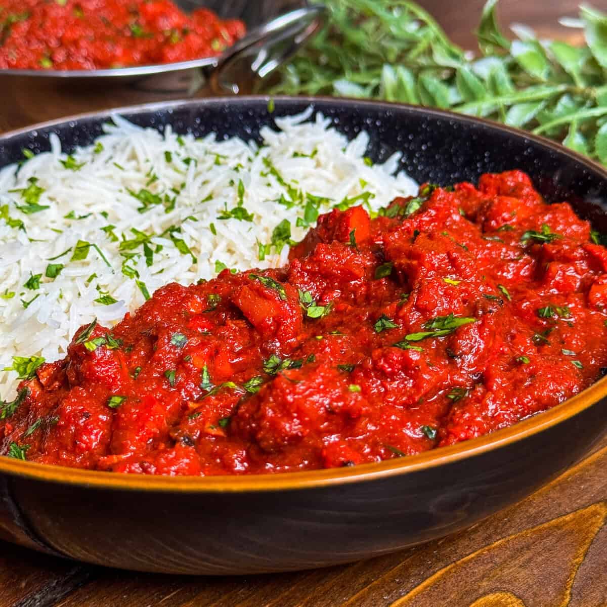 A bowl of white rice garnished with herbs sits next to a serving of vibrant red curry, also topped with fresh herbs. A small sprig of greens and another bowl of curry are in the background on a wooden surface.