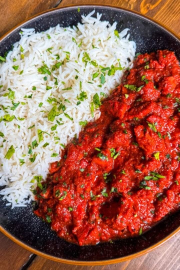 A black plate with white rice garnished with chopped herbs on one side and a vibrant red curry or stew, also topped with herbs, on the other side, set on a wooden table.