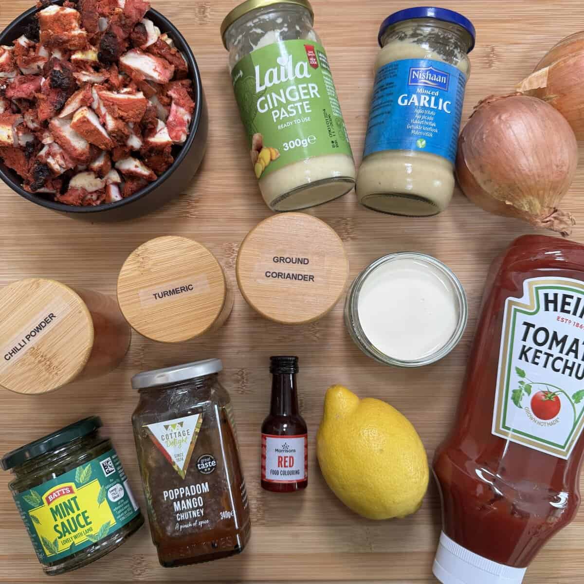 A flat lay of various cooking ingredients on a wooden surface, including chopped meat, jars of ginger paste and garlic, two onions, spice jars, cream, a lemon, mint sauce, mango chutney, red food coloring, and tomato ketchup.