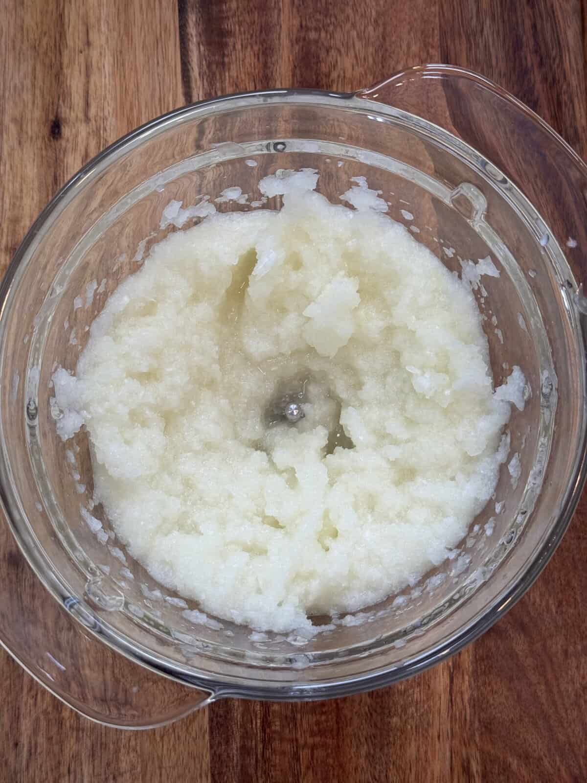 A glass bowl filled with finely blended or grated white onions sits on a wooden surface. The mixture appears moist and slightly chunky.