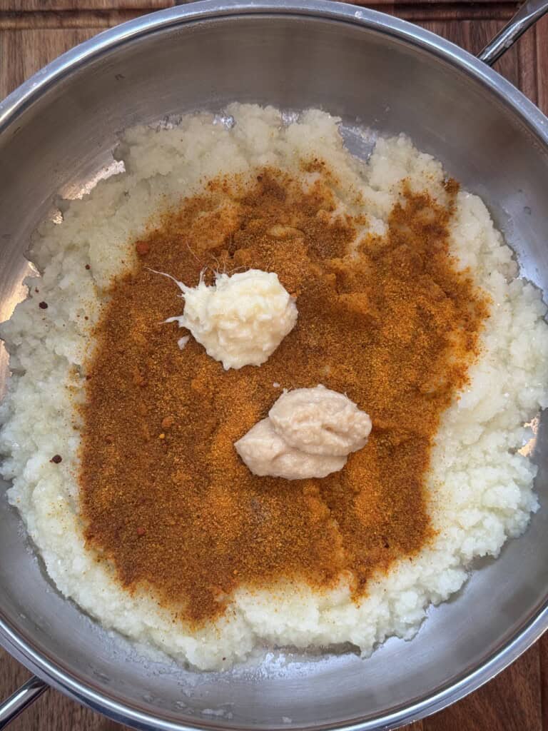 A metal pan contains cooked, mashed onions topped with orange-brown spices, a dollop of minced garlic, and a dollop of ginger paste, ready to be mixed together.