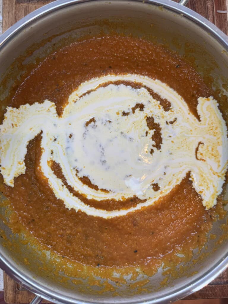 A pot of orange soup, likely tomato or carrot, with a swirl of cream added on top before being stirred in. The soup rests on a wooden surface.
