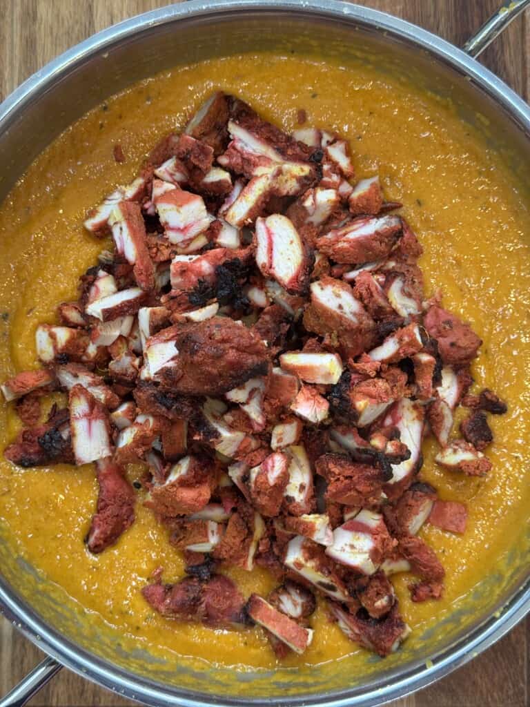 A stainless steel pan filled with bright orange curry sauce, topped with sliced pieces of tandoori chicken. The dish sits on a wooden surface.