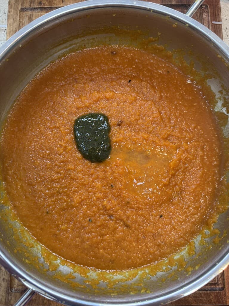 A pot filled with thick orange sauce or soup, with a dollop of dark green paste on top, sits on a wooden surface. The sauce appears freshly stirred and slightly textured.