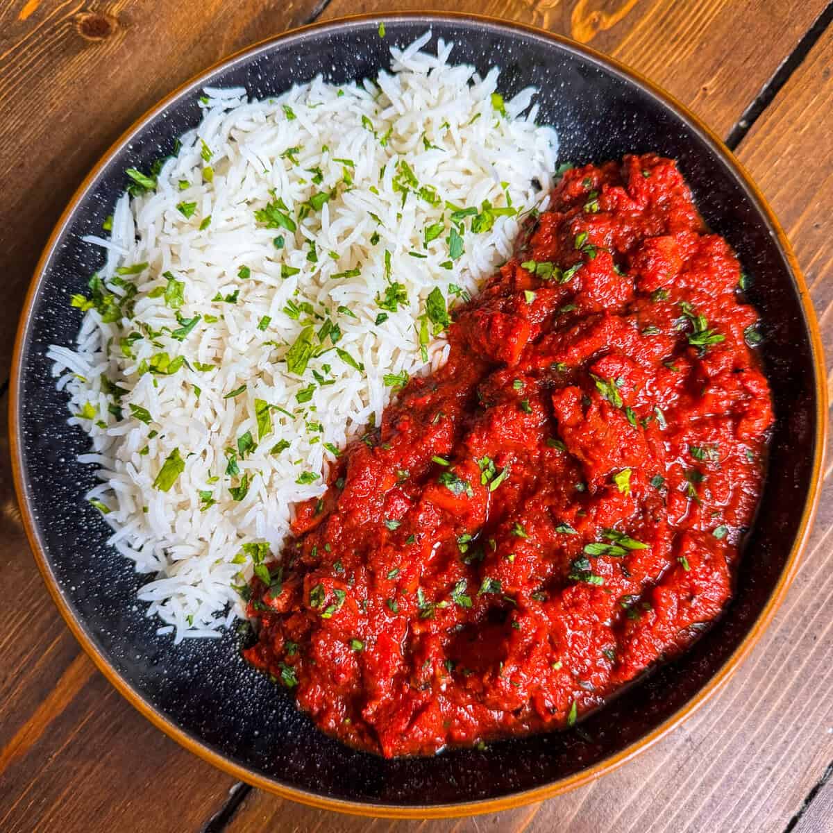 A black plate with white rice garnished with chopped herbs on one side and a vibrant red curry or stew, also topped with herbs, on the other side, set on a wooden table.