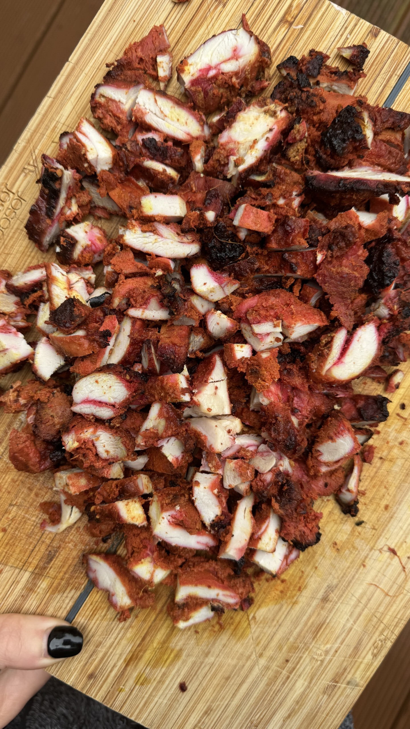 Chopped grilled meat with a reddish seasoning and charred edges is spread on a wooden cutting board. A hand with black nail polish holds one side of the board.