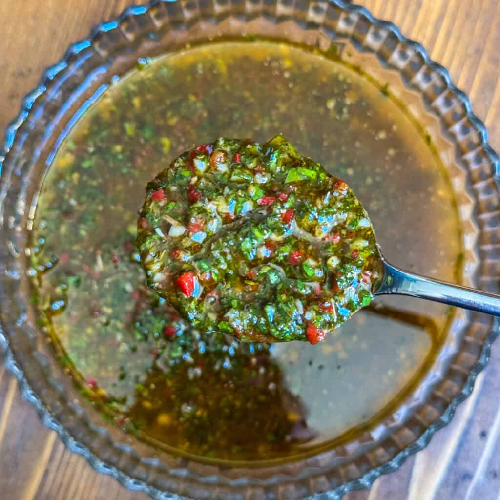 A close-up of a spoon holding chimichurri sauce above a glass bowl, showing a mixture of chopped herbs, red peppers, and oil on a wooden surface.