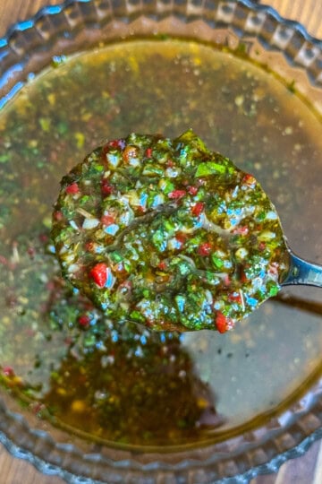 A close-up of a spoon holding chimichurri sauce above a glass bowl, showing a mixture of chopped herbs, red peppers, and oil on a wooden surface.