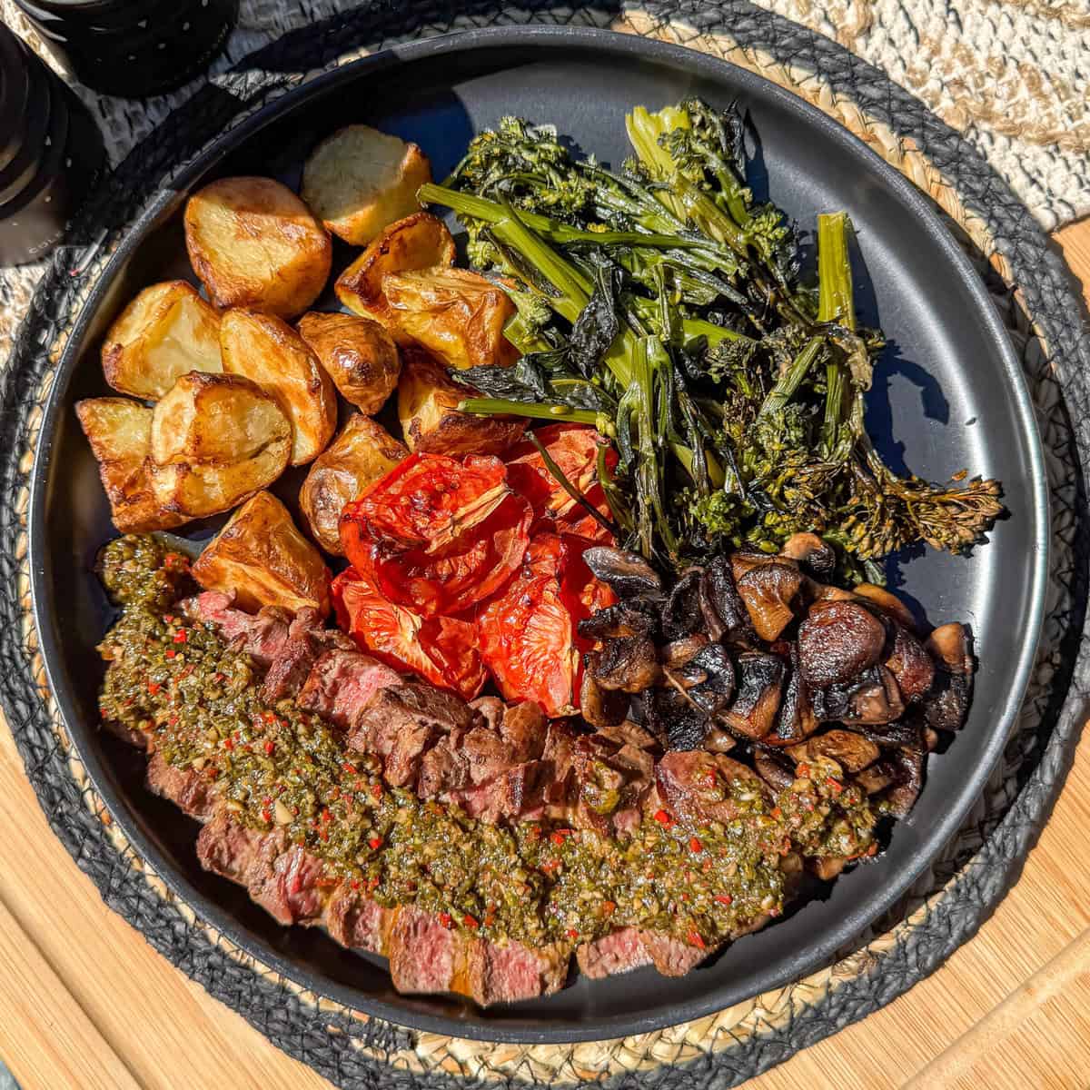 A black plate with sliced steak topped with chimichurri, roasted potatoes, broccolini, sautéed mushrooms, and roasted tomatoes, arranged in sections. The plate sits on a woven mat on a wooden surface.