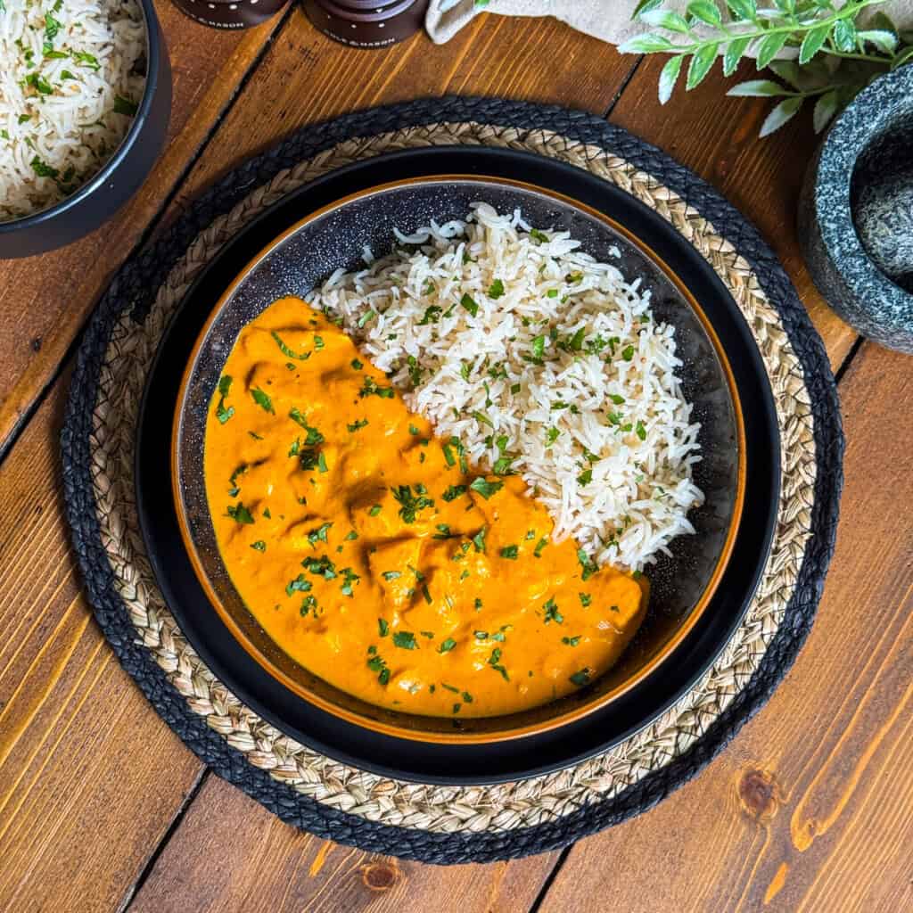A bowl of creamy orange curry with chunks of meat or tofu, garnished with herbs, served alongside white basmati rice. The bowl sits on a woven placemat atop a wooden table.