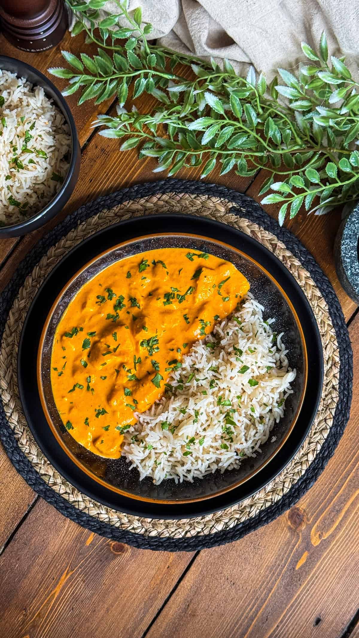 A black bowl contains white rice and creamy orange curry, garnished with chopped herbs. The bowl sits on a decorative mat, with a small bowl of rice and green leaves nearby on a wooden table.