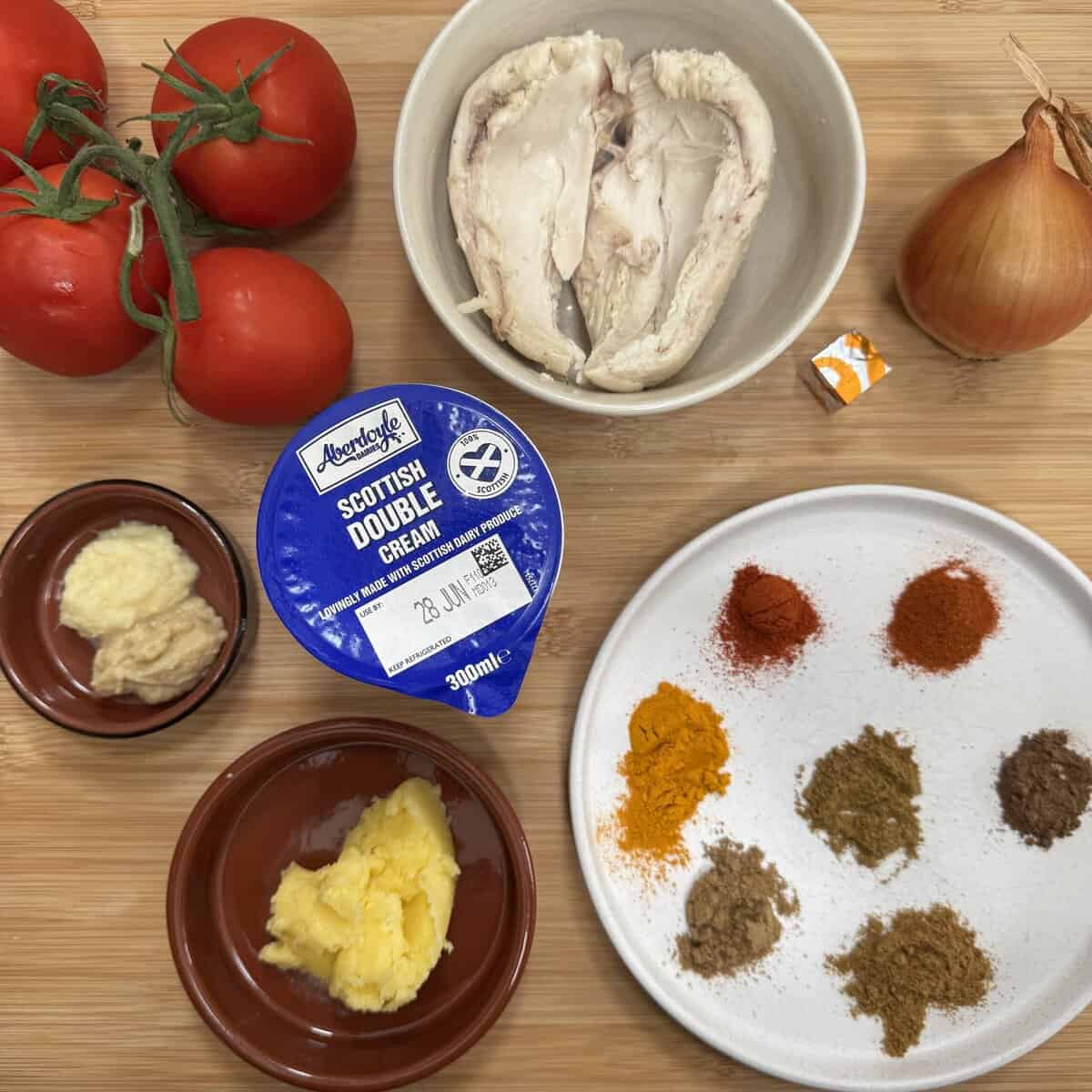 A wooden surface displays raw chicken in a bowl, three tomatoes, an onion, a chicken stock cube, a pot of Scottish double cream, ginger and garlic pastes, ghee, and small piles of various ground spices.